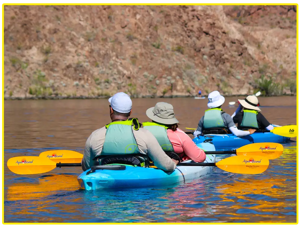 A row of tandem kayakers paddling together through brown canyon walls on a sunny day.