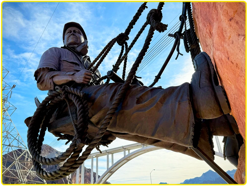 Bronze statue of a Hoover Dam worker suspended by ropes on a canyon wall, symbolizing the historic engineering of the Boulder Dam and Colorado River. The sculpture is set against the Black Canyon and desert landscape near Lake Mead, with the Hoover Dam overlook, powerlines, and the iconic Mike O’Callaghan–Pat Tillman Memorial Bridge in the background. This tribute honors Nevada’s history, the construction of the Hoover Dam, and its role in shaping the Southwest.