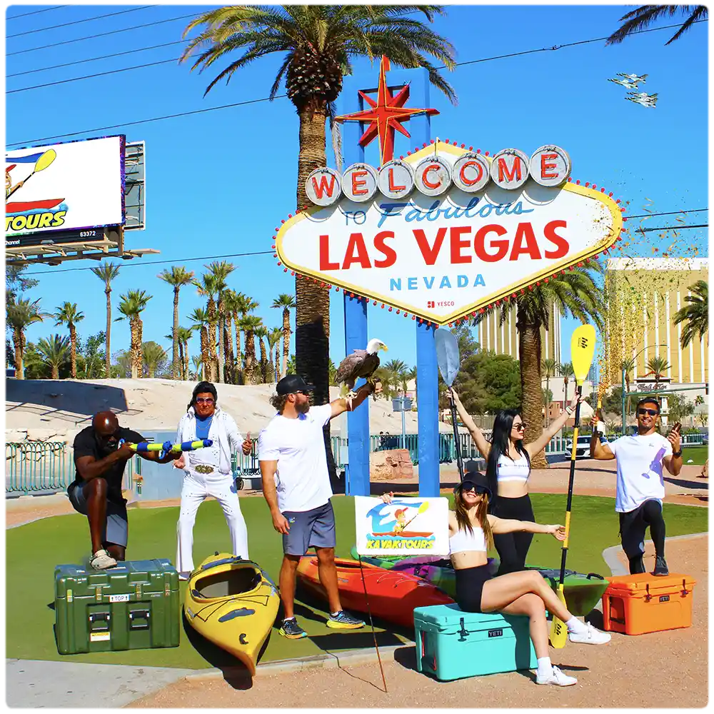 EZ Kayak Tours team posing with kayaks and paddles at the Welcome to Fabulous Las Vegas sign, promoting scenic guided kayaking tours on the Colorado River near Black Canyon and Emerald Cove.